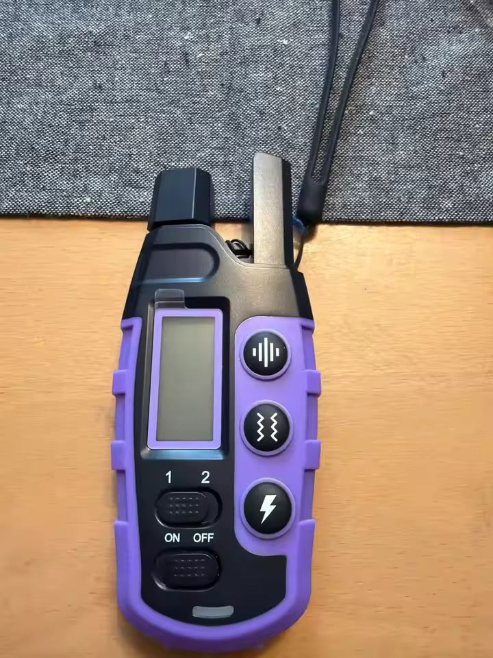 A handheld electronic device with a purple and black rugged housing, featuring a small screen, two buttons, and a wrist strap, resting on a wooden surface with a gray textured fabric background.