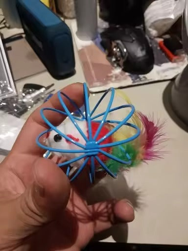 A hand holding a small toy with a blue spider-like wire frame, a white plush face, and colorful feathers in pink, yellow, green, and blue, against a cluttered desk background.